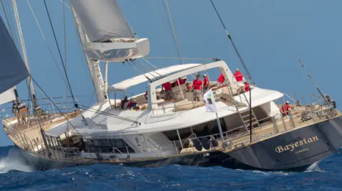 Perini Navi Press Office A photo of the Bayesian yacht pictured sailing. Several crew members in red tops can be seen. The ship's hull is a navy blue colour while it has one white sail and one blue sail.