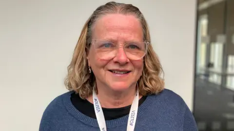 Shariqua Ahmed/BBC Head and shoulders image of Heather Skibsted. She has shoulder length light brown hair and is wearing clear rounded glasses. She is wearing a blue jumper with a black top underneath and a white lanyard.