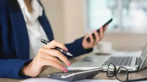 Close up image of a professional woman sitting at a computer making calculations and looking at her smartphone. She is wearing a blue blazer and an open neck white blouse. (Stock image)