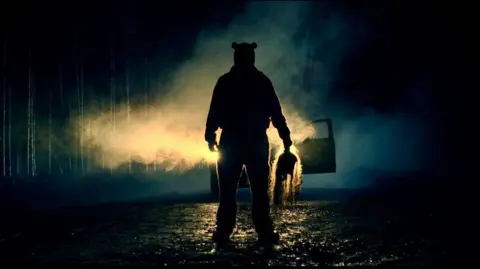 A silhouette of a figure in front of car headlights, at night. He is holding something ominous in his hand. 
