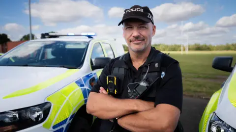 Lincolnshire Police DC Aaron Flint stands wearing police uniform in front of a police vehicle.