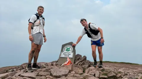 Pritchard Brothers The twon men from the above picture, standing on top of a pile of rocks. A marker shows they are on Pen y Fan. They are wearing shorts and trainers with white t-shirts and are carrying rucksacks.