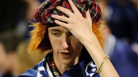A young man in a tartan see you jimmy hat holds his head in despair, with a gold wristband and blue flag draped over his shoulders.