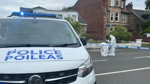 BBC A police van and police tape on a road, with two people in forensics outfits behind the tape working away.