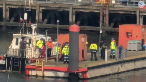 Supplied Rescued crew members wearing yellow high visibility jackets and blue helmets are seen disembarking from ships at Grimsby