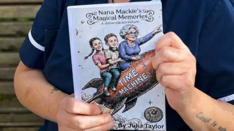 Julia Taylor Julia holding her book, Nana Mackie's Magical Memories: A Dementia Adventure. On the cover, there is a drawing of a rocket with the words "time machine" written on it. Sat on the rocket is an elderly woman who is pointing forward, and two children - a boy and a girl - sat behind her.
