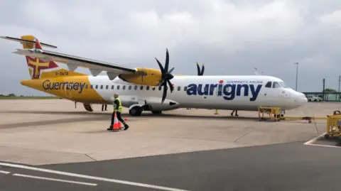 BBC A yellow and white Aurigny plane on a runway in Guernsey with a person wearing high vis carrying traffic cones walking beside the plane on an overcast day