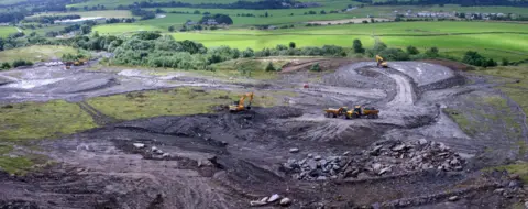 Steve Chettle Arts UK An opencast coal mine with trucks and diggers on site and green hills in the background