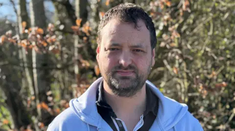 Quinn looking at the camera. He has brown hair and is wearing a grey zip up hoodie with a black top with a collar under it.