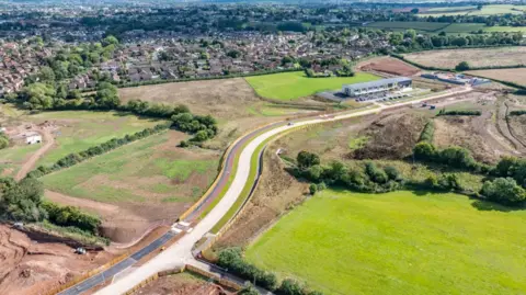 Barrett Photographers An aerial view of the partially competed Orchard Grove development on the outskirts of Taunton. A new primary school stands in the background, with a long road snaking through fields towards it. 