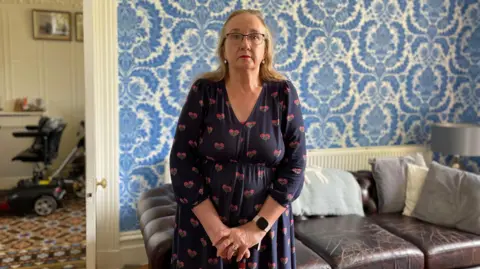 Joanne Greenhalgh is pictured wearing a navy dress patterned with flowers and leaning on a walking stick.