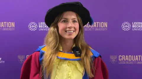 A woman in a cap and gown stands in front of a purple background as she receives her honorary doctorate.