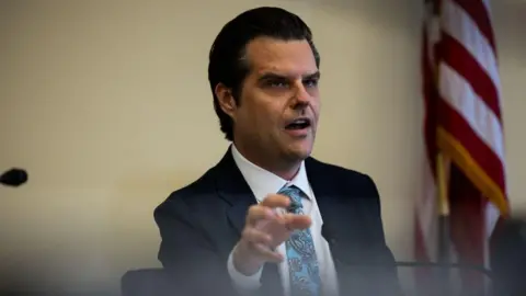 Getty Images Matt Gaetz, wearing a suit, gestures with his first hand while sitting and speaking at a microphone during a hearing 