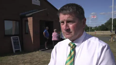 A man with brown hair is wearing a white shirt with a gold and green striped tie. He is standing in front of a red brick village hall. There is a flag pole in the background with a union jack on it