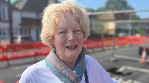 Tom Jackson/BBC Sarah Payne has short blonde hair and is wearing a purple cotton T-shirt and a green patterned scarf. She is standing in front of a sinkhole which has a metal fence around it.