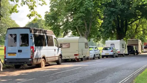A road running through a green space. A line of vans and caravans are parked along it as well as a couple of cars. 