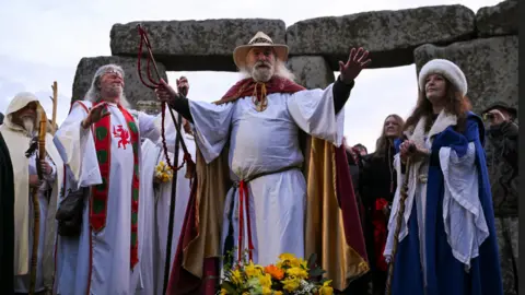 Men and women in robes look spiritual and stand in front of stones. A man in front raises his arms.