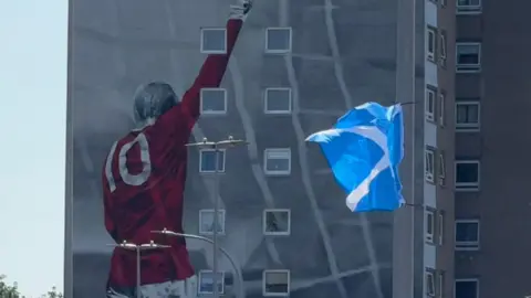 A mural of a Aberdeen FC player Denis Law with his arm raised in the air while wearing a red shirt with the number 10 on it is painted on the side of a tower block. A saltire which has been tied to a lamppost waves in the wind in front of the tower block. 