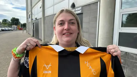 Matt Dean/BBC Alice Blyth holding her new shirt aloft. She has blonde hair and brightly-coloured bangles on her right wrist.