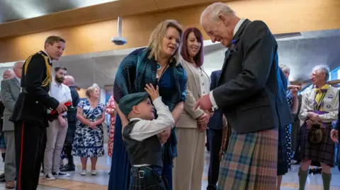 PA Media Four-year-old Gregor Corse holds up his his hand to King Charles with his fingers raised. They are both wearing traditional Scottish outfits including kilts. Gregor is also wearing a flat cap. Also pictured is Gregor's mum who is wearing a blue and green tartan shawl.
