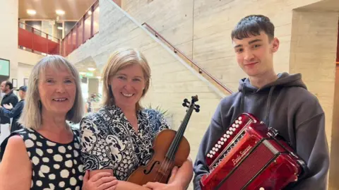A women with short blonde hair in a black and white dress, next to a women with a blue and white top holding a fiddle. On the far right a boy with a grey jumper holding an accordion.