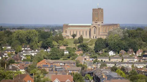 Getty Images Guildford Cathedral is the largest building in a landscape that also includes houses and trees and a blue sky behind it. The cathedral is a modern-looking one with a square tower rising at its middle and long thin windows along its visible side