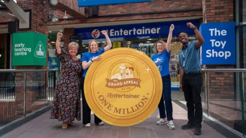 The Entertainer Four people are standing outside 'The Entertainer' toy shop in Cabot Circus, punching the air. Two are wearing 'The Entertainer' t-shirts. They are holding a giant cut-out of a gold coin which says 'one million'