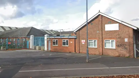 Google Maps A brick building. There are four parking spaces and a lamppost outside and blue metal fencing around parts of the building.