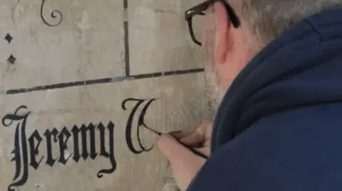 A signwriter adds Jeremy Wheeldon's name to a stone wall using a thinly tipped paintbrush and black paint.