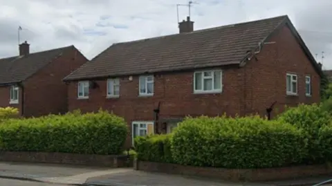A brick two-storey house with six white UPVC windows on the first floor and a similar window and door visible on the ground floor. There is a similar property next door with one visible UPVC window on the first floor. The properties have one brick chimney each. There is a brick wall and large bush surrounding the property.
