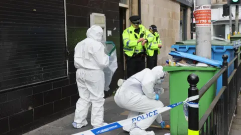 Colin Mearns/ The Herald and Glasgow Times A street scene: two forensics officers in white overalls, masks, blue gloves are in the street, with their backs to camera. One on the left holds an open plastic evidence bag, the other on the right is crouching near a green refuse bin. Behind them are two police officers in hi-vis jackets, standing either side of a building entrance. Police tape is in the foreground, attached to black railings.