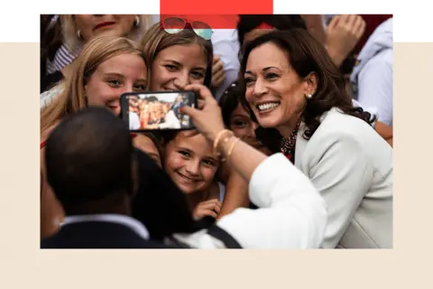 Getty Images Kamala Harris in a white jacket having her photo taken on a phone. She is posing with a group of young women and what appears to be a child