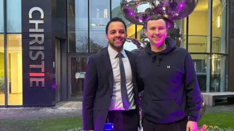 Sam Birchall Sam Birch with Dr Aziz Gulamhusein - a man who looks slightly older than him and he has black hair and a short black beard. Dr Gulamhusein wears a suit with a dark patterned tie and striped shirt. Sam wears a black hoodie with a small white logo. Both smile outside the front of the Christie building