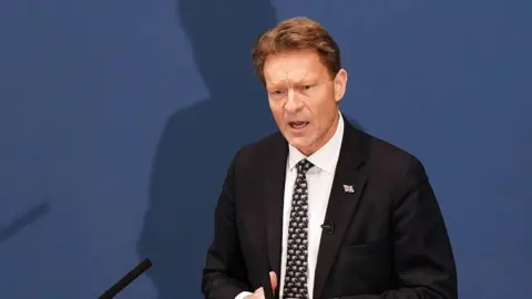 Richard Tice delivering his speech. He's wearing a dark suit, white tie and dark tie with pattern, standing in front of a blue display.