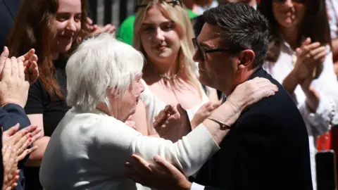 PA Media Andy Burham, side-on, is holding the elbow of his mother Eileen, who has grey hair and is wearing a white jumper. They are about to embrace.