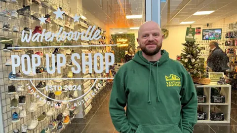 Matt Shaul, in a green Vintage Trainers branded hoodie stood outside the front of the new store which has "Waterlooville pop up shop Est 2024 Independent Authentic" printed on the glass outside. Behind the glass inside the store a man is shopping and there are walls full of shoes.