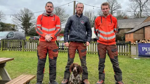 BBC Three men and a dog are stood in the picture. Andre and Bobby are wearing orange, high visibility jackets, while Jake is wearing a dark navy coat. Trevor is a spaniel and is in the centre of the photo. 