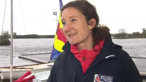 Hannah Snellgrove giving a TV interview. She has long brown hair, tied back. She is wearing a navy blue jacked with a British Sailing Team logo on the chest. It's a grey day and behind her is a lake with some boats moored on a pontoon.