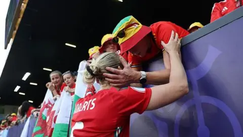 Rhiannon Roberts with her back to the camera. She is at the audience stand holding on to a woman's arm. The woman is holding on to Rhiannon's face and is consoling her while other fans to her left look on. 