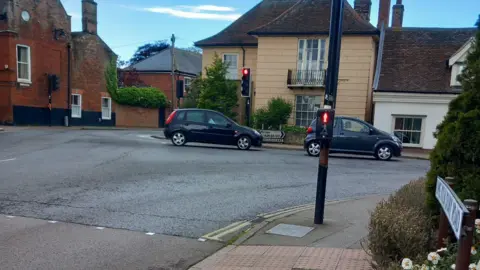 Gail Ratcliffe A road junction where there is a traffic light crossing. Two cars are ahead travelling along a road to the right. 