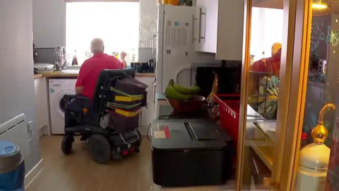 Tom in his electric wheelchair in his kitchen, with his back to the camera. A display cabinet can be seen to the left, with the kitchen sink, washing machine and counter in front.
