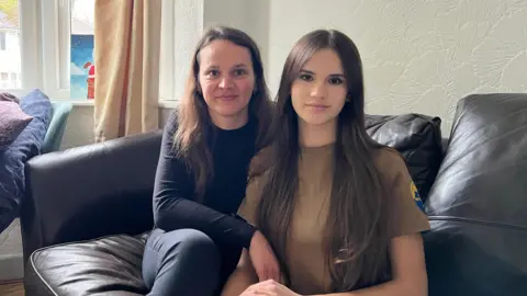 A woman with brown hair dressed all in black is sat on a black leather sofa next to a young woman who is wearing a brown T-shirt.