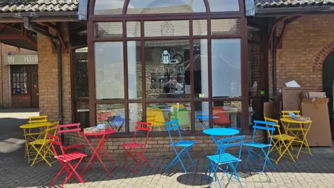 Project 16:15 A cafe, with colourful tables and chairs outside, boxed to the right and a large wooden window, with Cafe 16:15 written on the glass.