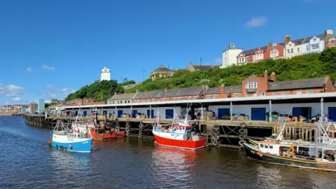 The Fish Quay in North Shields. Several small but brightly coloured fishing boats are moored in the harbour. A small blue boat is sailing in to dock. Wooden docks line the water and stone houses stand behind them.