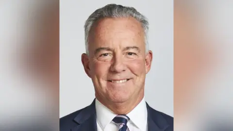 Sean Bell A man with grey hair smiling at the camera in a professional headshot. He is wearing a dark navy suit, white shirt and a striped tie in navy and lilac. The background is plain light grey.