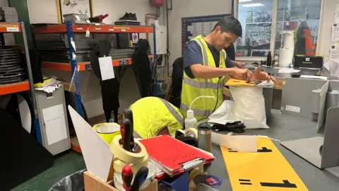 Oliver Whitfield Miocic/BBC A man wearing a hi-vis yellow jacket and a dark navy shirt is doing some handiwork on a table.