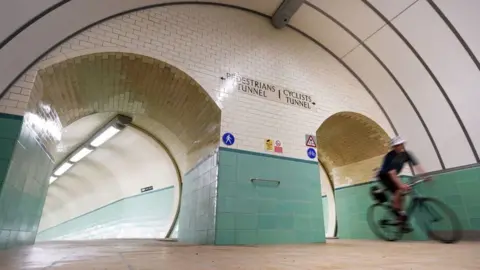 A cyclist rides through the Tyne Pedestrian and Cyclist Tunnel. He is wearing a white helmet and black clothing. The tunnel has blue-green tiles on the lower section and white tiles above. A pedestrian tunnel runs alongside the cycling one.