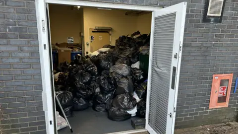 A huge mound of rubbish in a store room. There are lots of black and grey bins and containers full of rubbish bags behind them. Piled up recycling is away in the corner. 