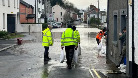 Riverside area road is flooded, people in high-vis carry sandbags to doors and by the river bridge. 