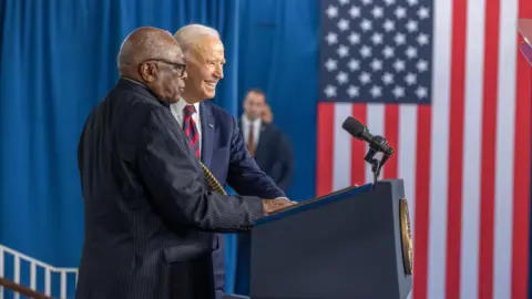 Jim Clyburn appears at a podium with Joe Biden, who is smiling, with an American flag in the background